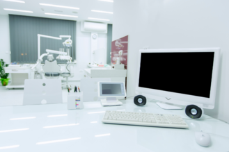 Modern dental office with a white desk holding a monitor, keyboard, mouse, speakers, and a digital device, possibly for patient check-in. In the background, a dental chair, overhead light, and instruments are visible, all within a bright, sterile environment featuring white walls and vertical blinds.