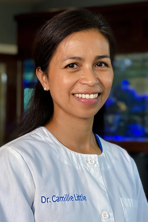 Dr. Camille Little wearing a white lab coat with her name embroidered in blue; background features soft blue lighting and a fish tank, suggesting a calming, professional environment.