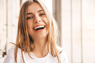 Smiling person with straight light brown hair in white shirt, standing in front of sunlit vertical window panels.